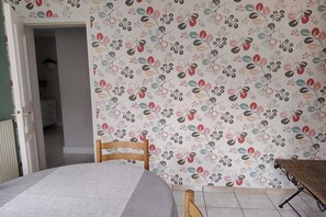 Une pièce avec un lit, une chaise en bois, un papier peint à motifs et une vue sur une autre pièce.
