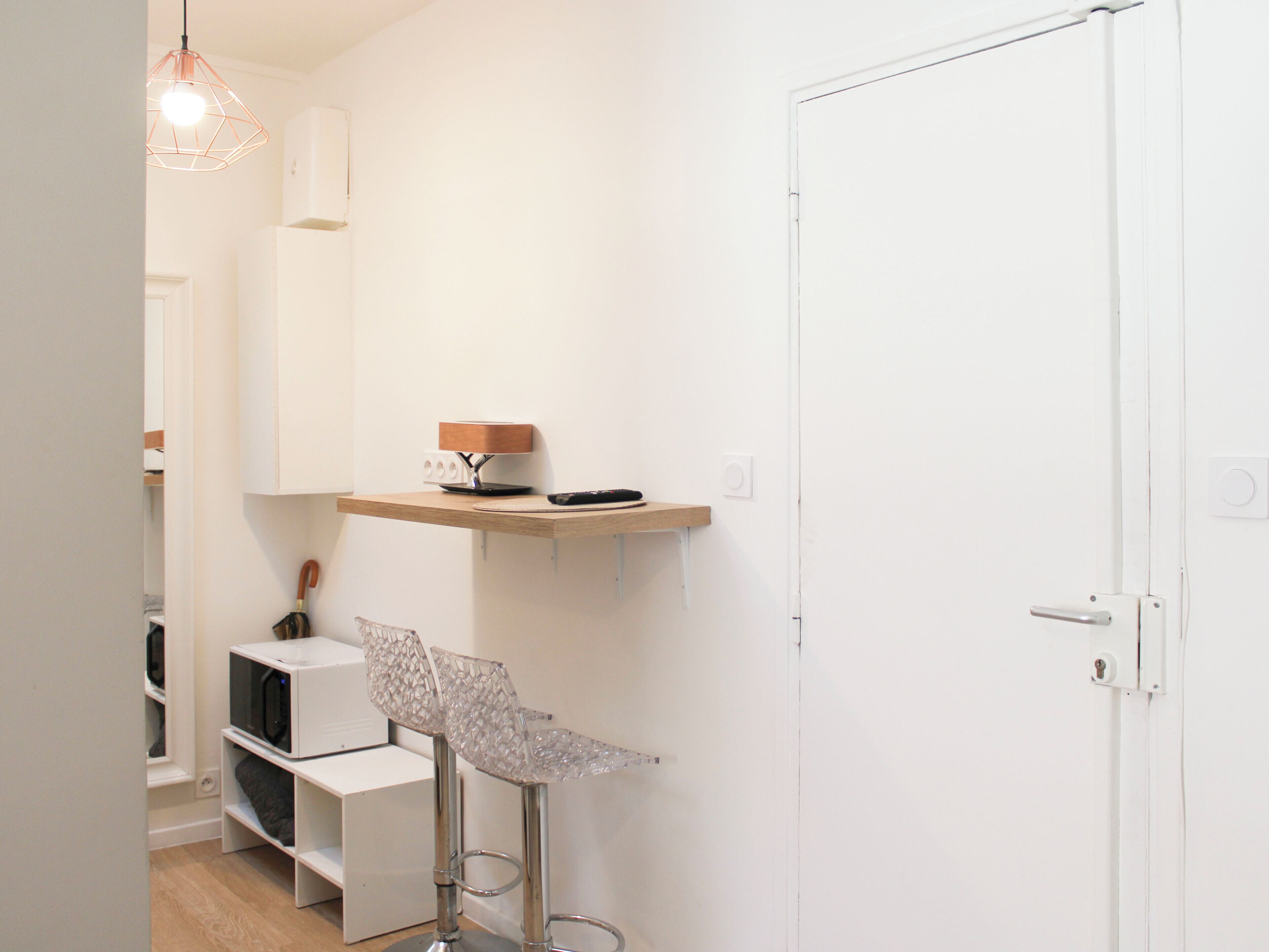 Une pièce minimaliste avec une porte blanche, un petit coin bar avec un tabouret, une étagère avec une lampe et un luminaire suspendu.