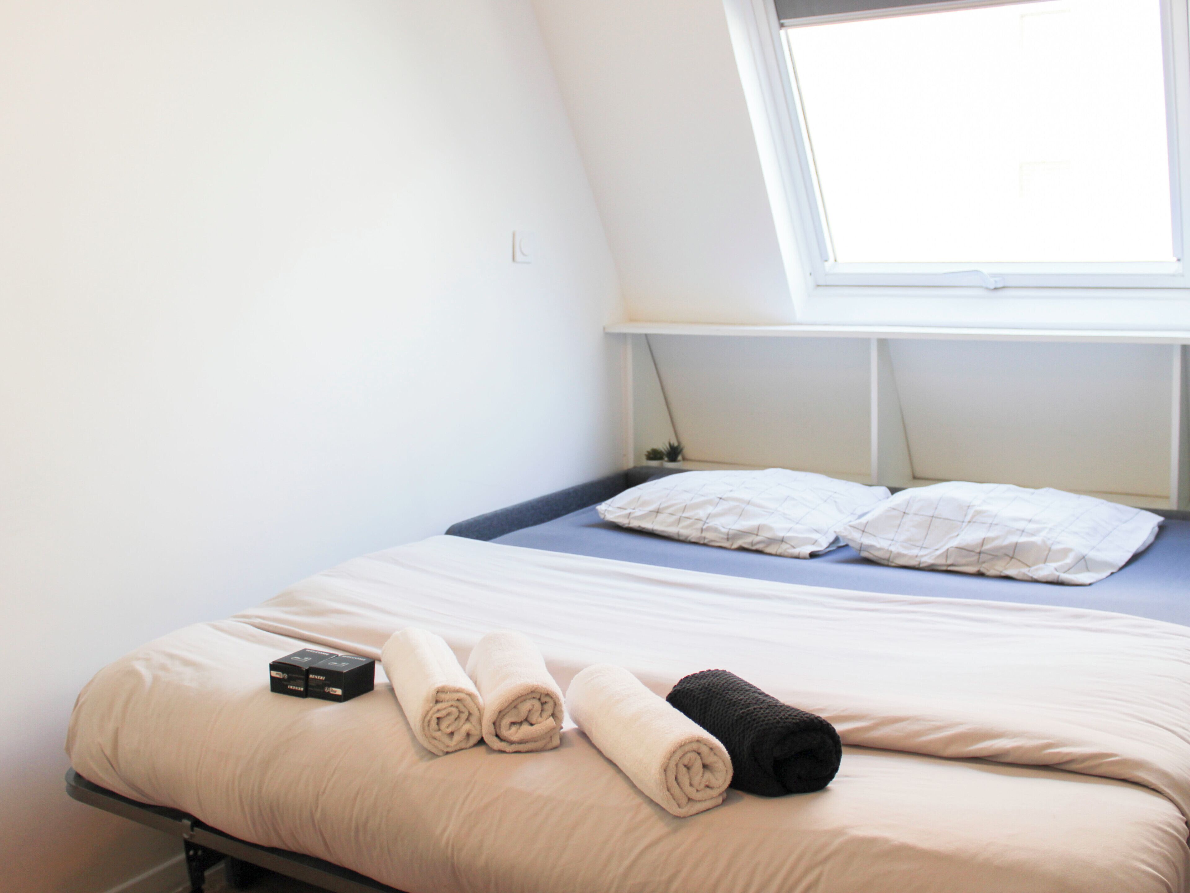 Een eenpersoonsbed met een wit-beige dekbedovertrek, twee opgerolde handdoeken en een zwart voorwerp op het bed.