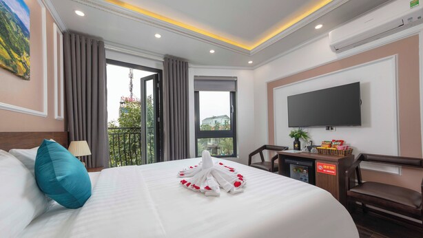 Een hotelkamer met een netjes opgemaakt bed, een flatscreen televisie aan de muur, een bureau met een computer en een kleine plant, en een balkon met uitzicht op de stad.