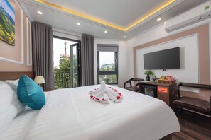 Une chambre d’hôtel avec un lit parfaitement fait, un téléviseur à écran plat fixé au mur, un bureau avec un ordinateur et une petite plante, et un balcon offrant une vue sur la ville.