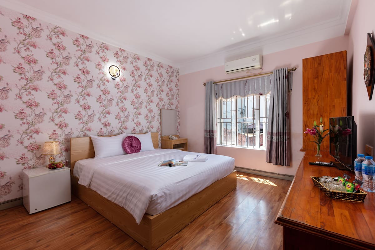 A bedroom with a wooden bed, a nightstand, a bedside table, a window with curtains, a wall-mounted air conditioner, and a wooden dresser with toiletries.