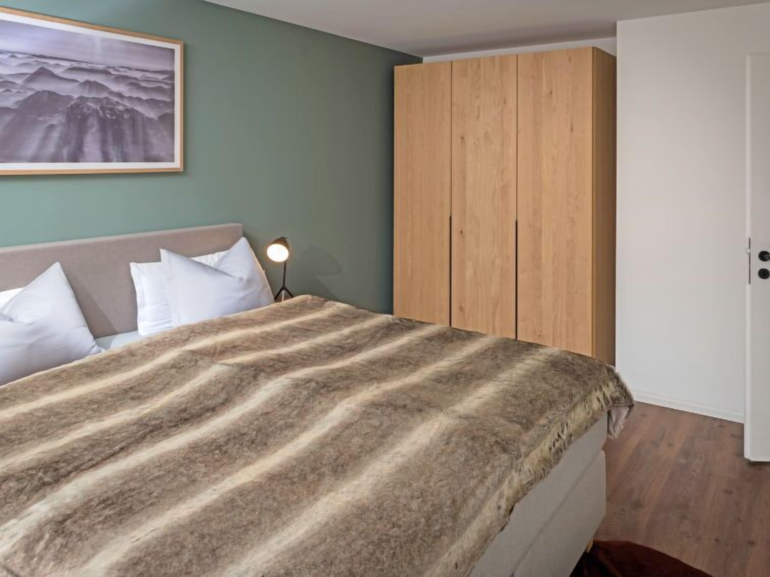A bedroom with a bed, a wooden wardrobe, a nightstand with a lamp, and a framed picture on the wall.