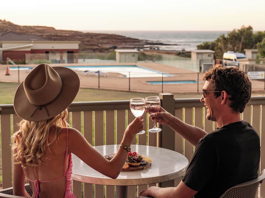 A couple seated at a round table on a wooden deck overlooking a pool and ocean view.