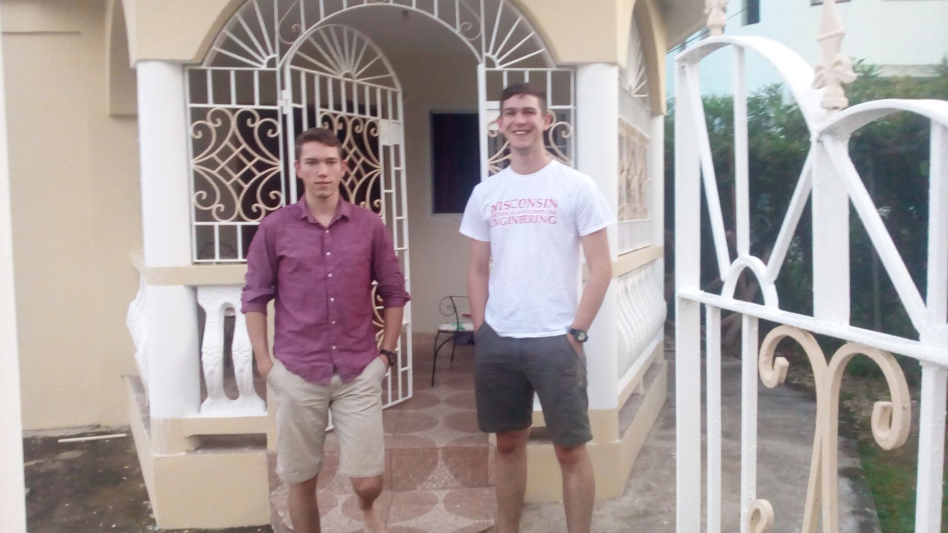 Deux hommes se tiennent devant un immeuble orné d’une grille décorative et doté d’un petit balcon.