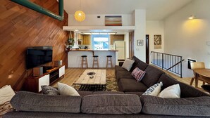 Ein modernes Wohnzimmer mit einer großen Couch, einem Couchtisch und einem Fernseher. Im Hintergrund befindet sich ein Küchenbereich mit einer Bar und Hockern.