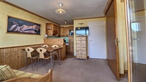 Een kleine keuken met houten kastjes, een eethoek met een tafel en stoelen, en een koelkast.