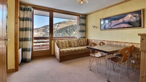 Een kamer met een houten bank, een eettafel met stoelen en een groot raam met uitzicht op bergen.