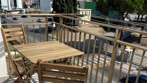 A balcony with a wooden table and chairs, a metal railing, and a view of the cityscape.