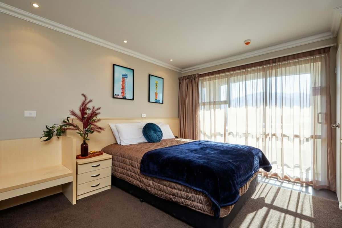 A hotel room with a bed, a bedside table, a lamp, and a window with curtains.