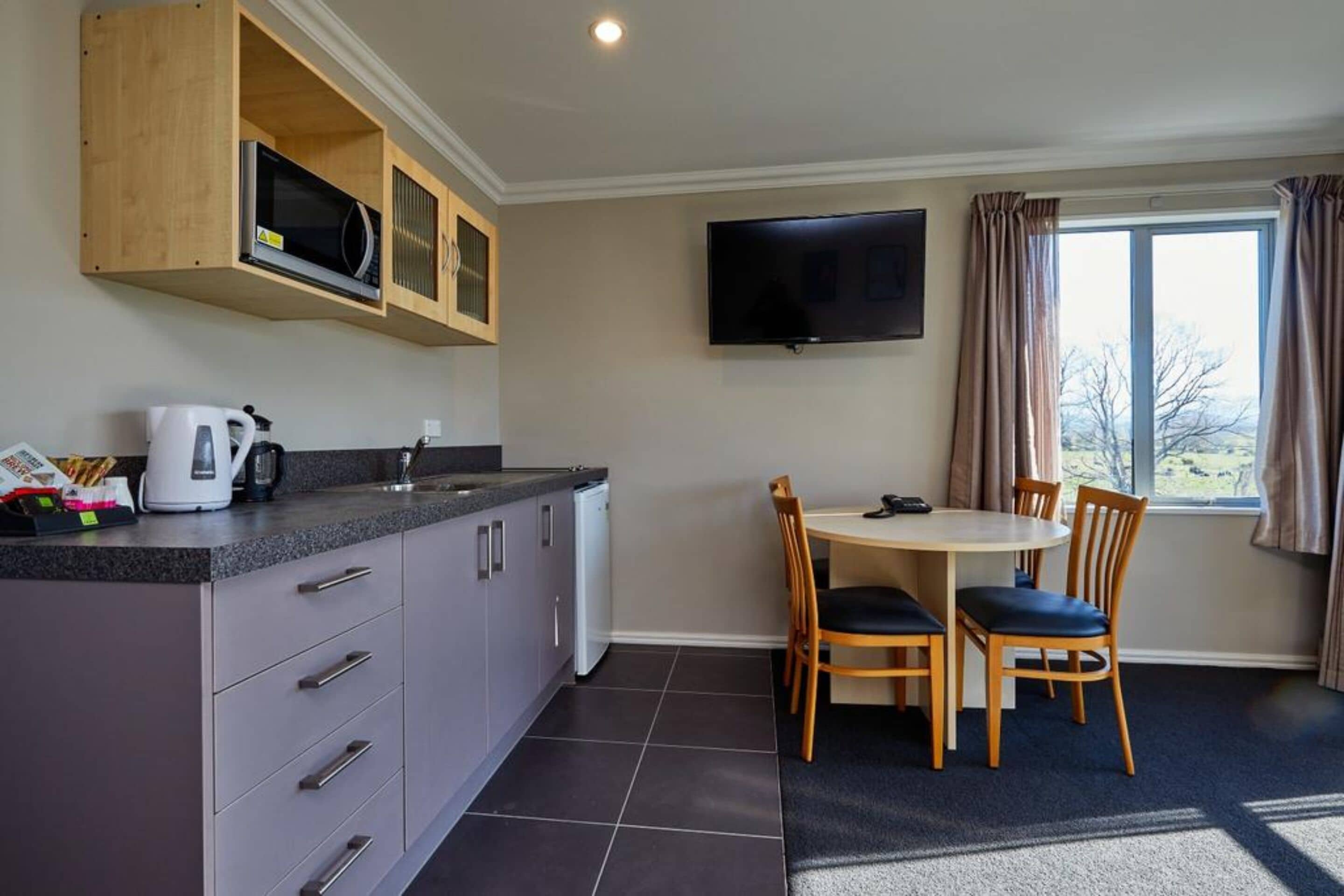 A compact kitchen with a microwave, kettle, and coffee maker, a dining area with a table and chairs, and a TV mounted on the wall.