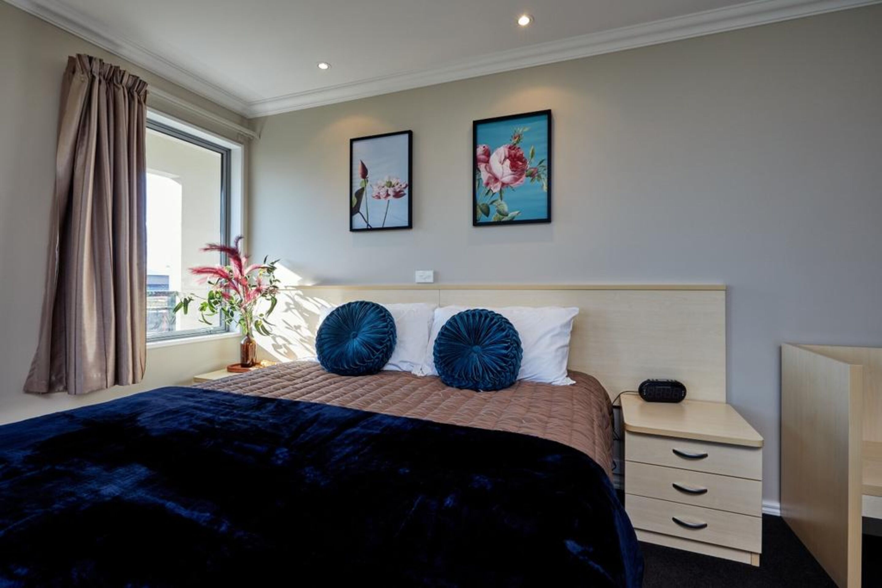 Habitación de hotel con una cama grande, una mesita de noche con lámpara, una ventana con cortinas y dos cuadros en la pared.