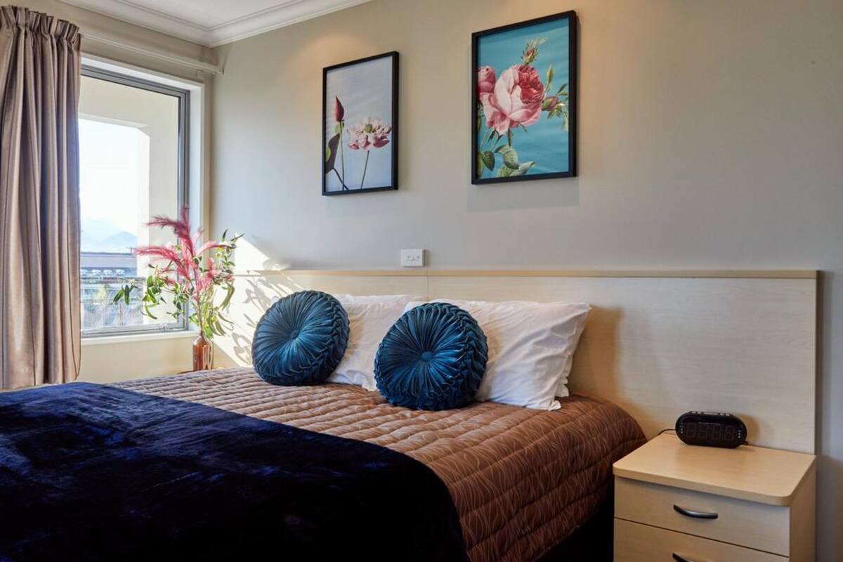 A hotel room with a bed, a nightstand, a window with curtains, and two framed floral paintings on the wall.