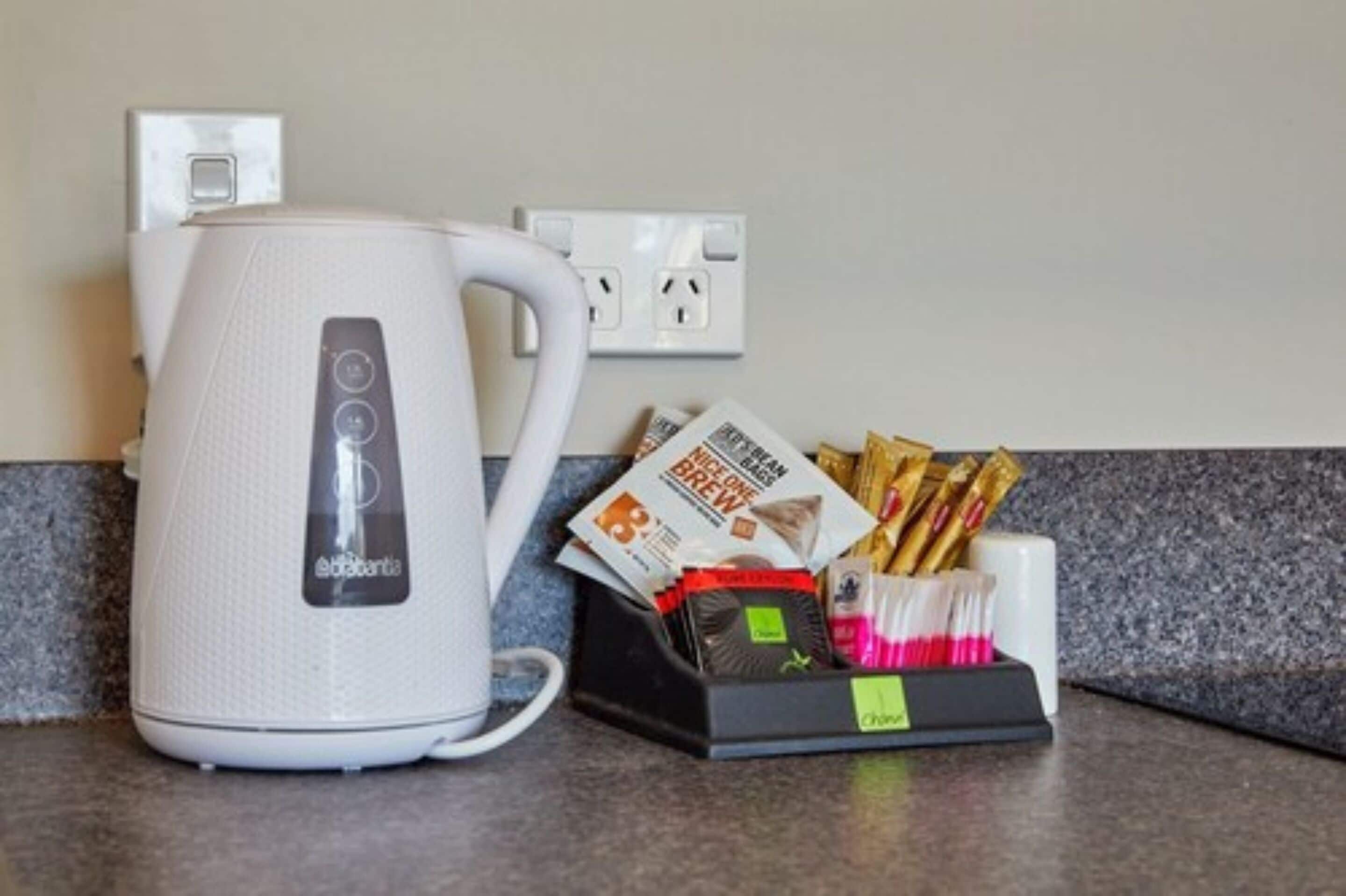 A white electric kettle on a kitchen counter with a coffee maker, a coffee pod holder, and a coffee grinder.