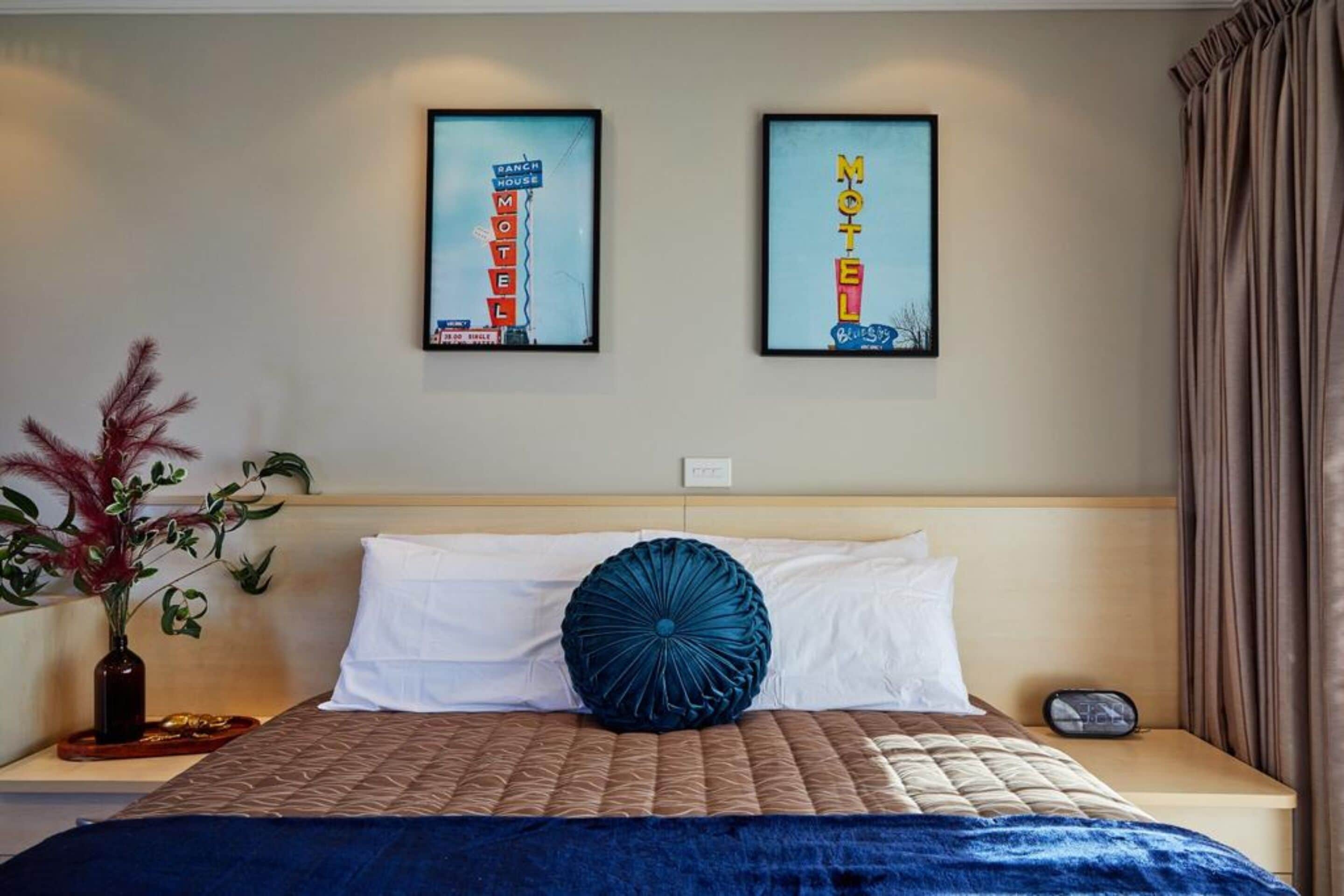 Et hotelværelse med en seng, et natbord, en vase med planter og to billeder i rammer på væggen.