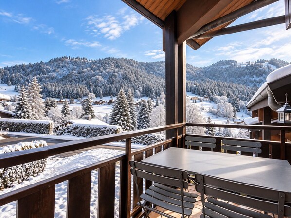 A wooden balcony with a table and chairs overlooking a snowy landscape with trees and houses.
