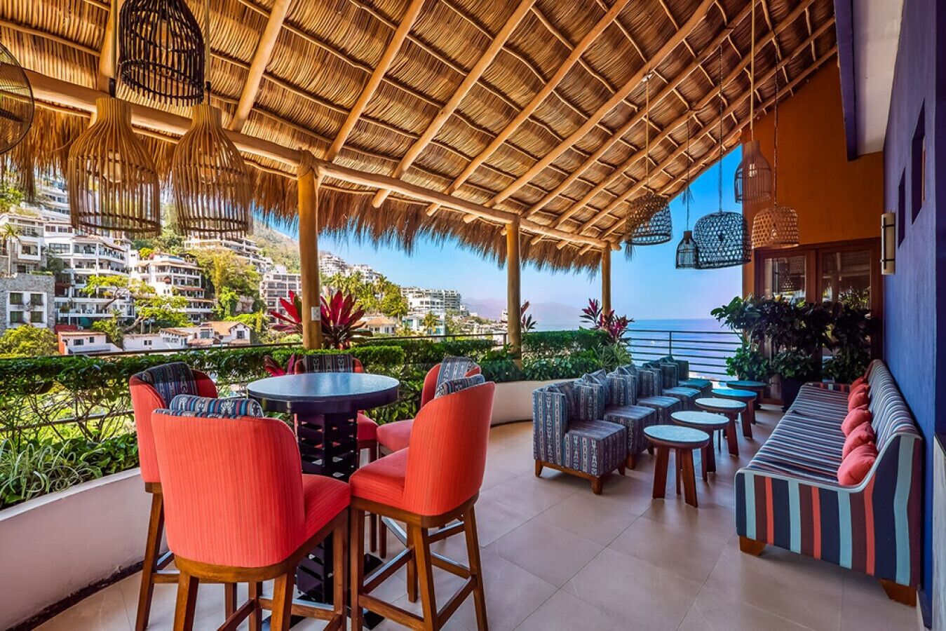 Un espacio al aire libre cubierto con mesas y sillas redondas, un sofá a rayas y vistas al mar y edificios al fondo.