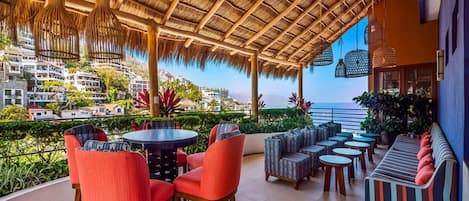 Un espacio al aire libre cubierto con mesas y sillas redondas, un sofá a rayas y vistas al mar y edificios al fondo.