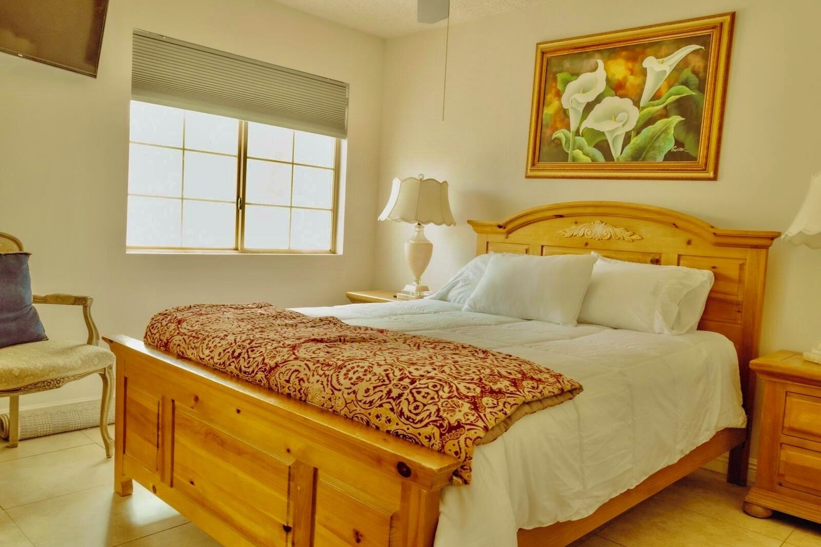 Une chambre avec un lit en bois, une fenêtre avec des stores, une chaise, une table de chevet avec une lampe et un tableau représentant des fleurs accroché au mur.
