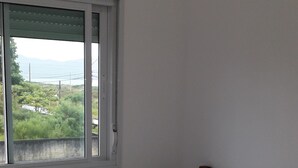 A single bed with a wooden headboard in a room with a window overlooking greenery and a distant view. - 04 - Holiday apartment - Morada Madruga (Praia do Sonho (Ens Brito))