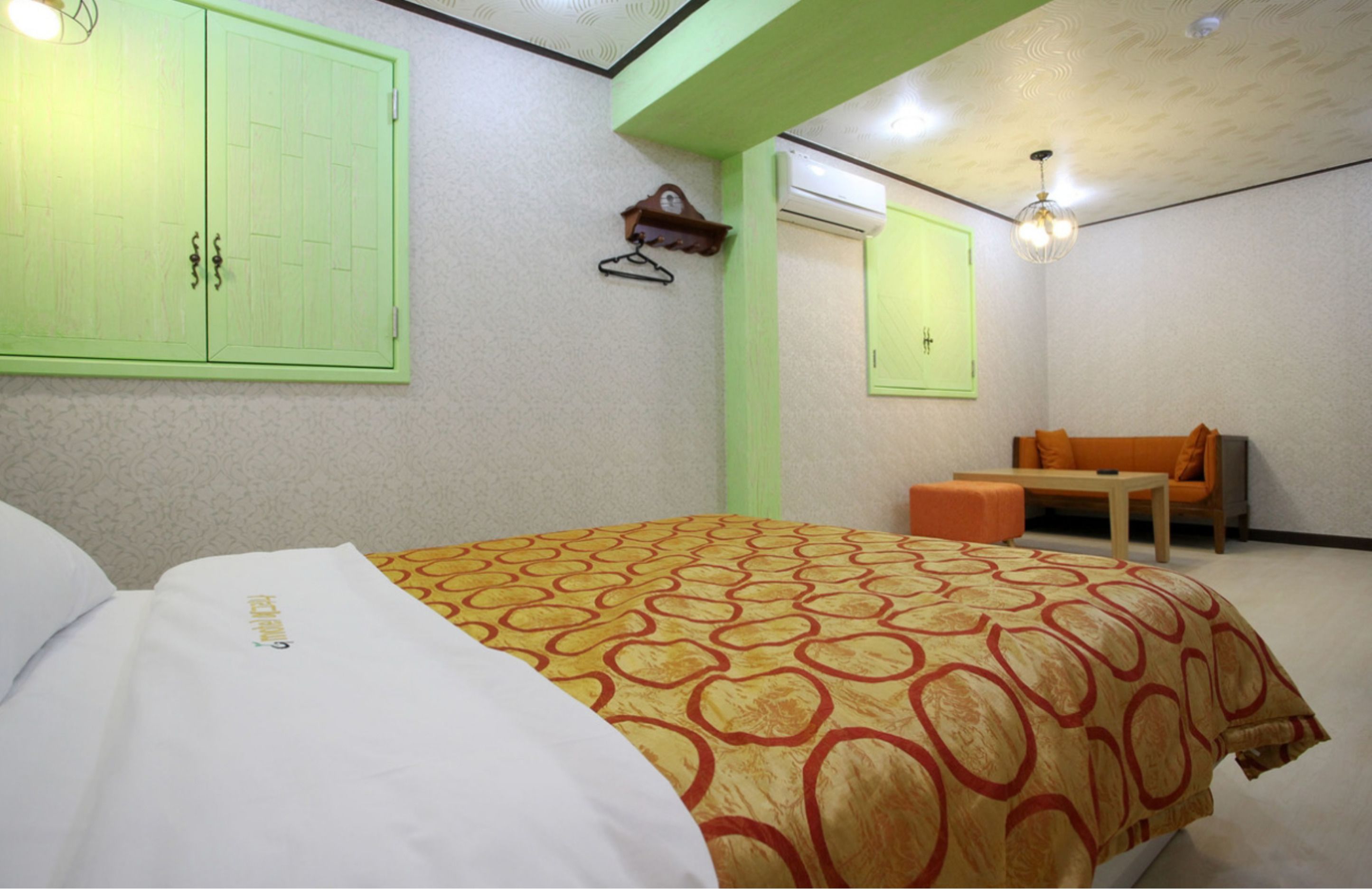 A hotel room with a bed, a chair, a small table, a wall-mounted air conditioner, and a light fixture.