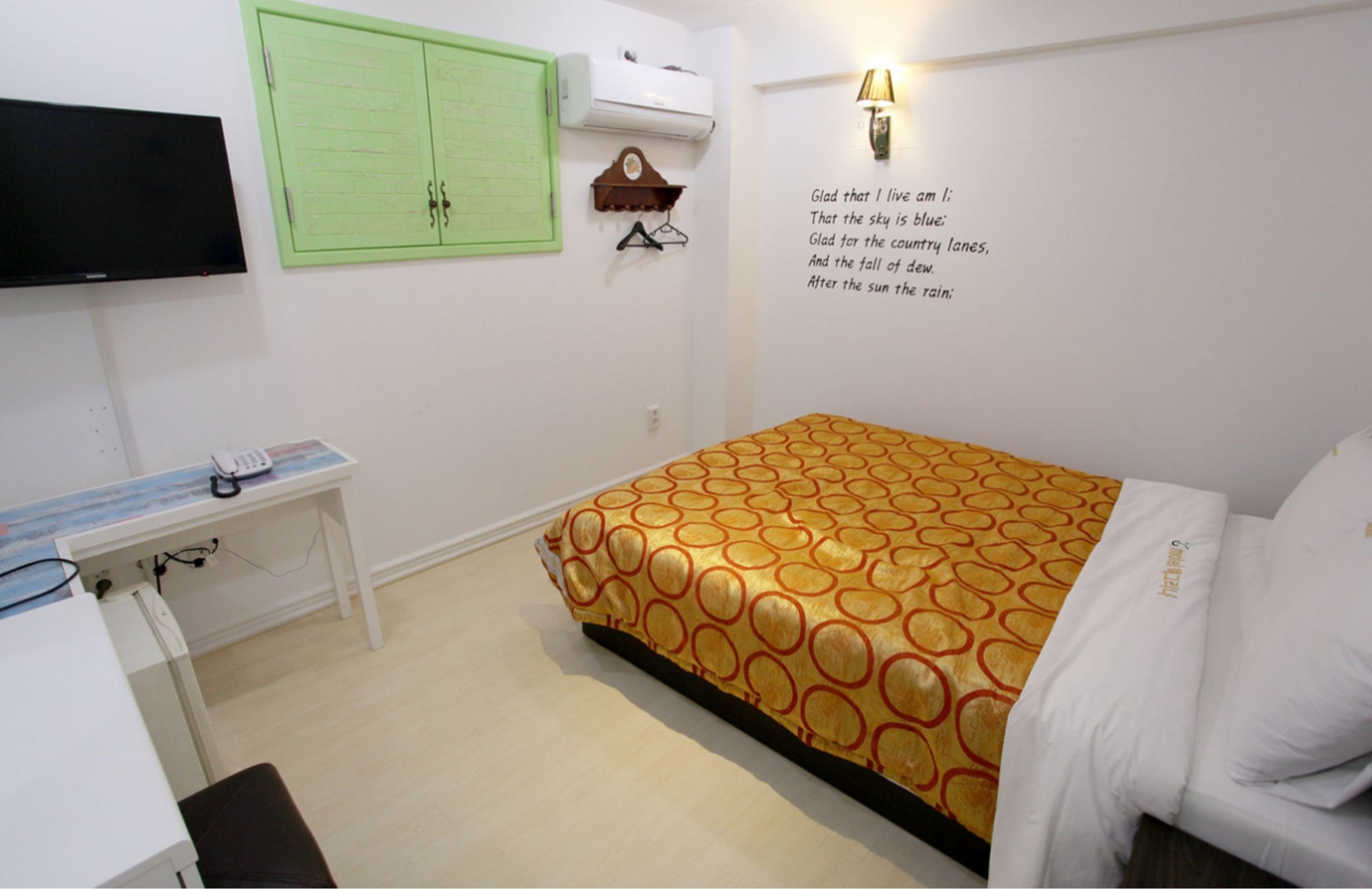 A bedroom with a bed, a desk, a television, a green cabinet, and a wall-mounted lamp.