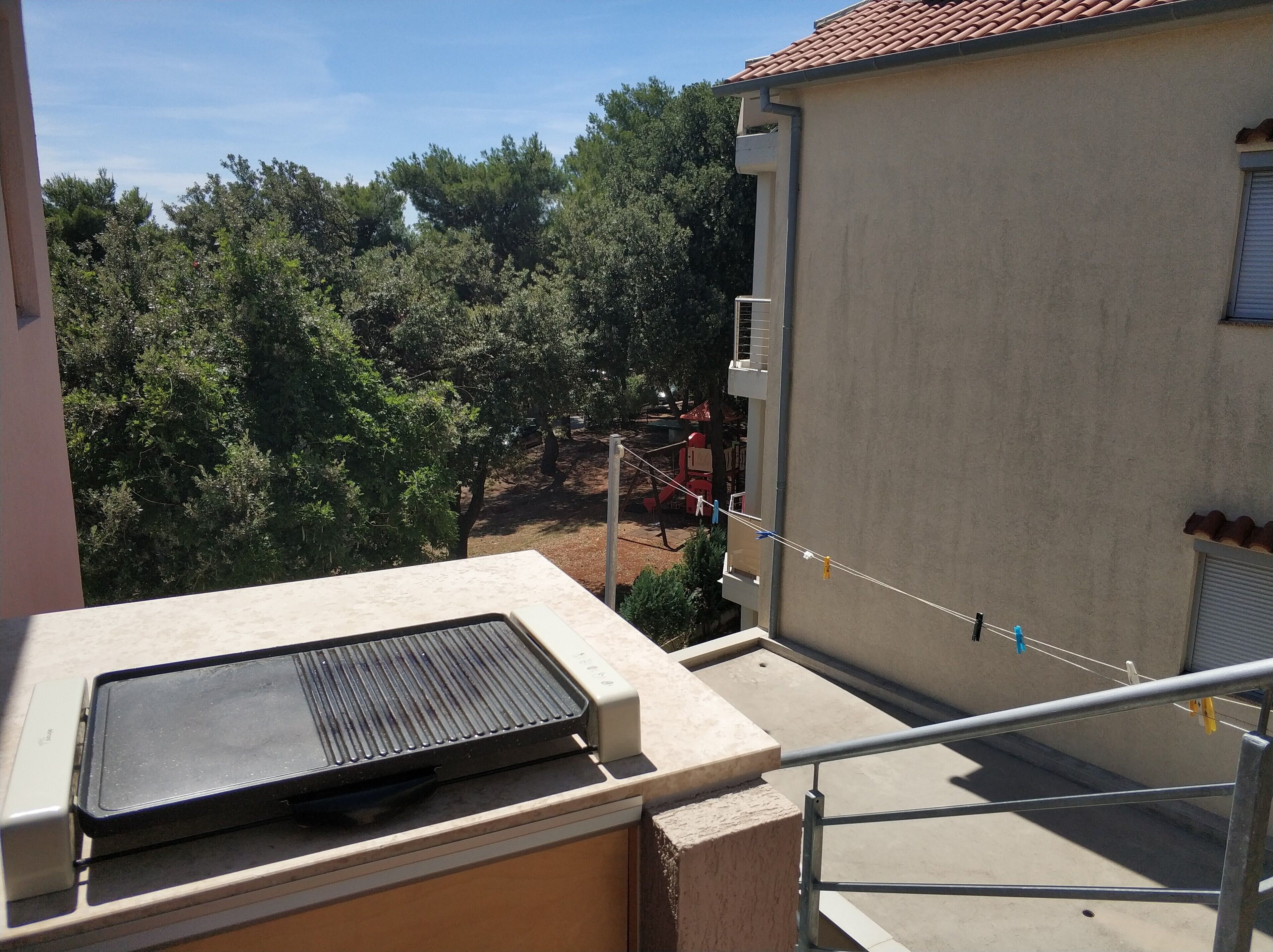 Un balcon avec un barbecue, une vue sur les arbres et une aire de jeux en arrière-plan.