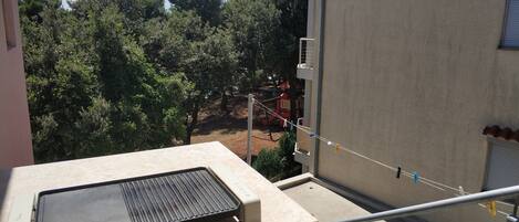 Un balcon avec un barbecue, une vue sur les arbres et une aire de jeux en arrière-plan.
