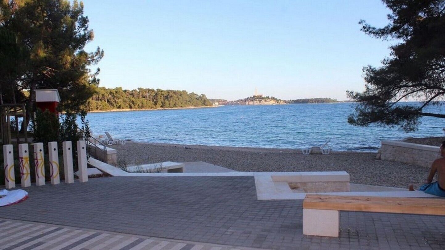 Ein Strand mit einer gepflasterten Promenade, Bänken und weiter Aussicht auf einen Hügel mit einem Turm.