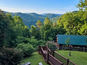 A cabin with a wooden deck and a green roof, surrounded by lush forest and mountains.