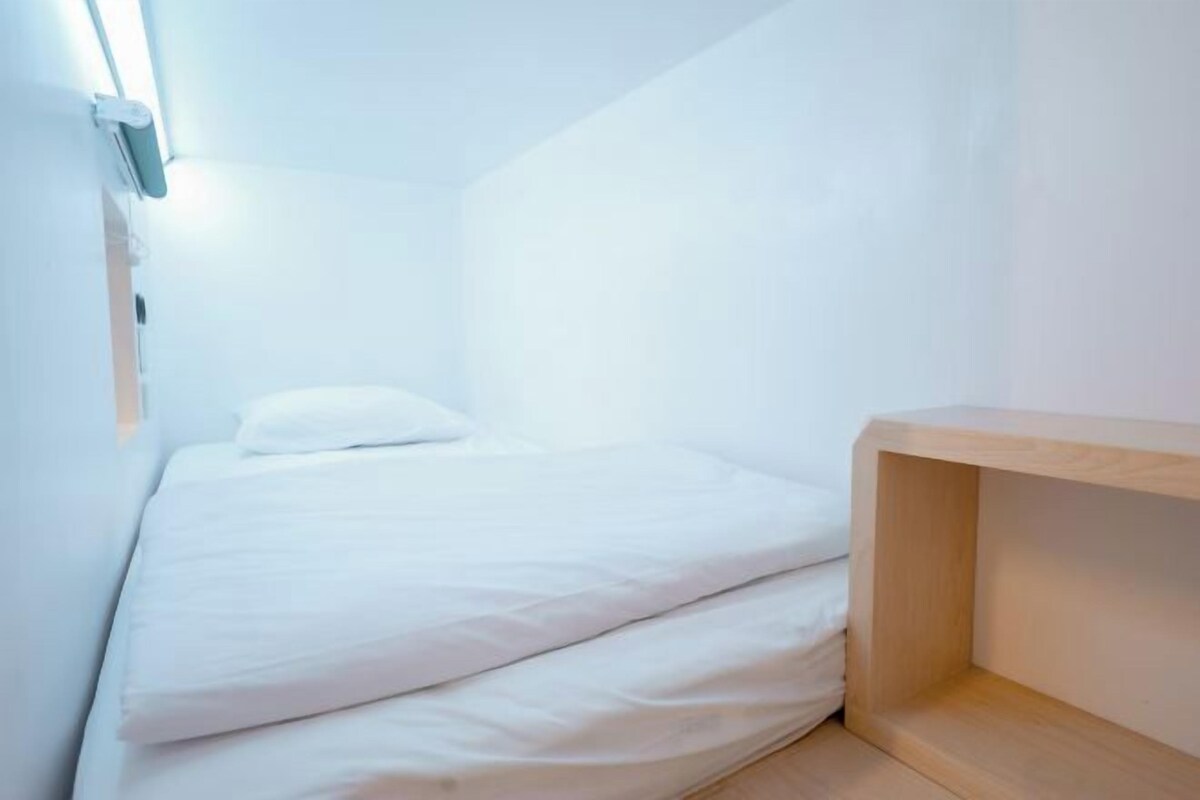 Una habitación pequeña con una cama individual, un mesita de noche de madera y una lámpara de pared.
