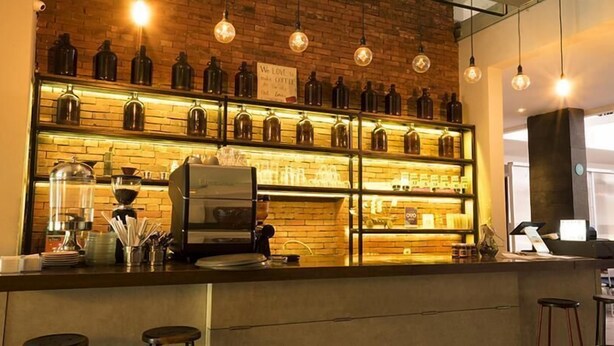 A bar area with a coffee machine, shelves of bottles, and hanging light bulbs.
