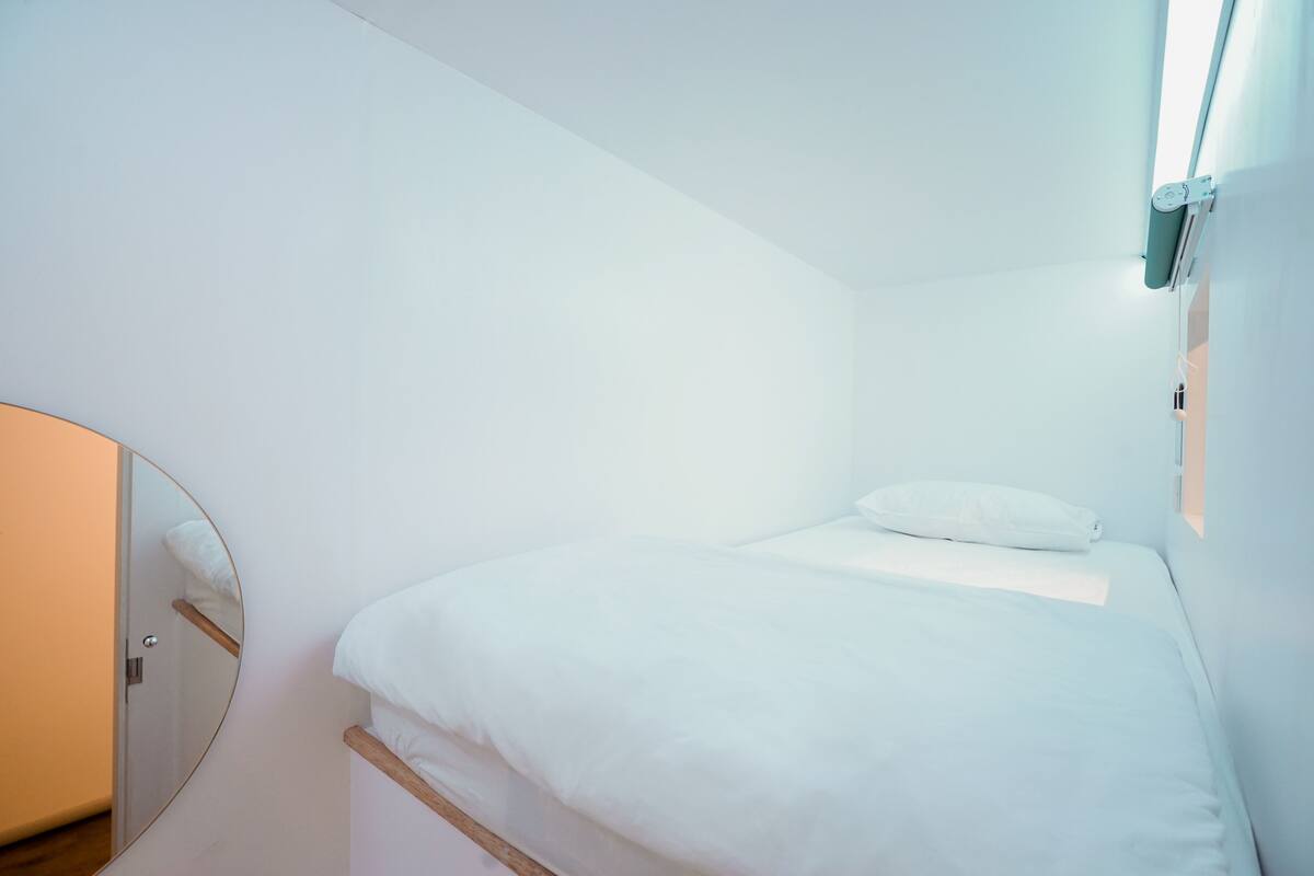 Een kleine, witte kamer met een eenpersoonsbed, een ronde spiegel aan de muur en een nachtkastje.