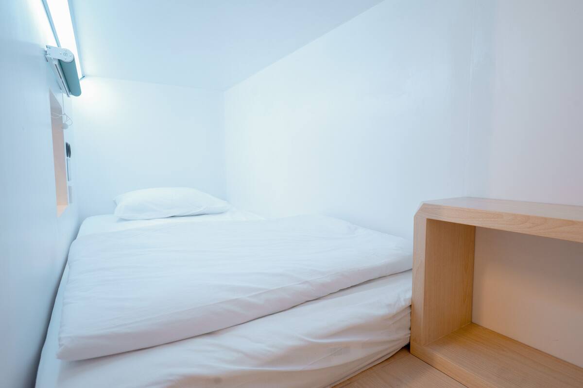 Een klein, netjes opgemaakt bed met witte lakens in een hoek slaapkamer, met een houten plankje en een wandlamp.
