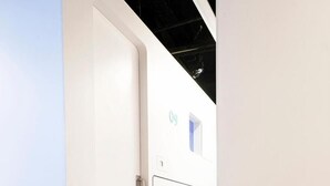 A corridor with white walls and wooden flooring. There are doors on the left and right, and a small window on the right wall.