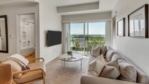 Una sala de estar moderna con sofá, sillón y mesa de centro. Un televisor está montado en la pared, y hay un balcón con vistas.