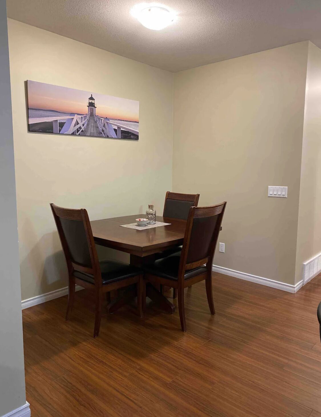 Un coin repas avec une table en bois et quatre chaises, un tableau représentant un pont accroché au mur et un luminaire au plafond.