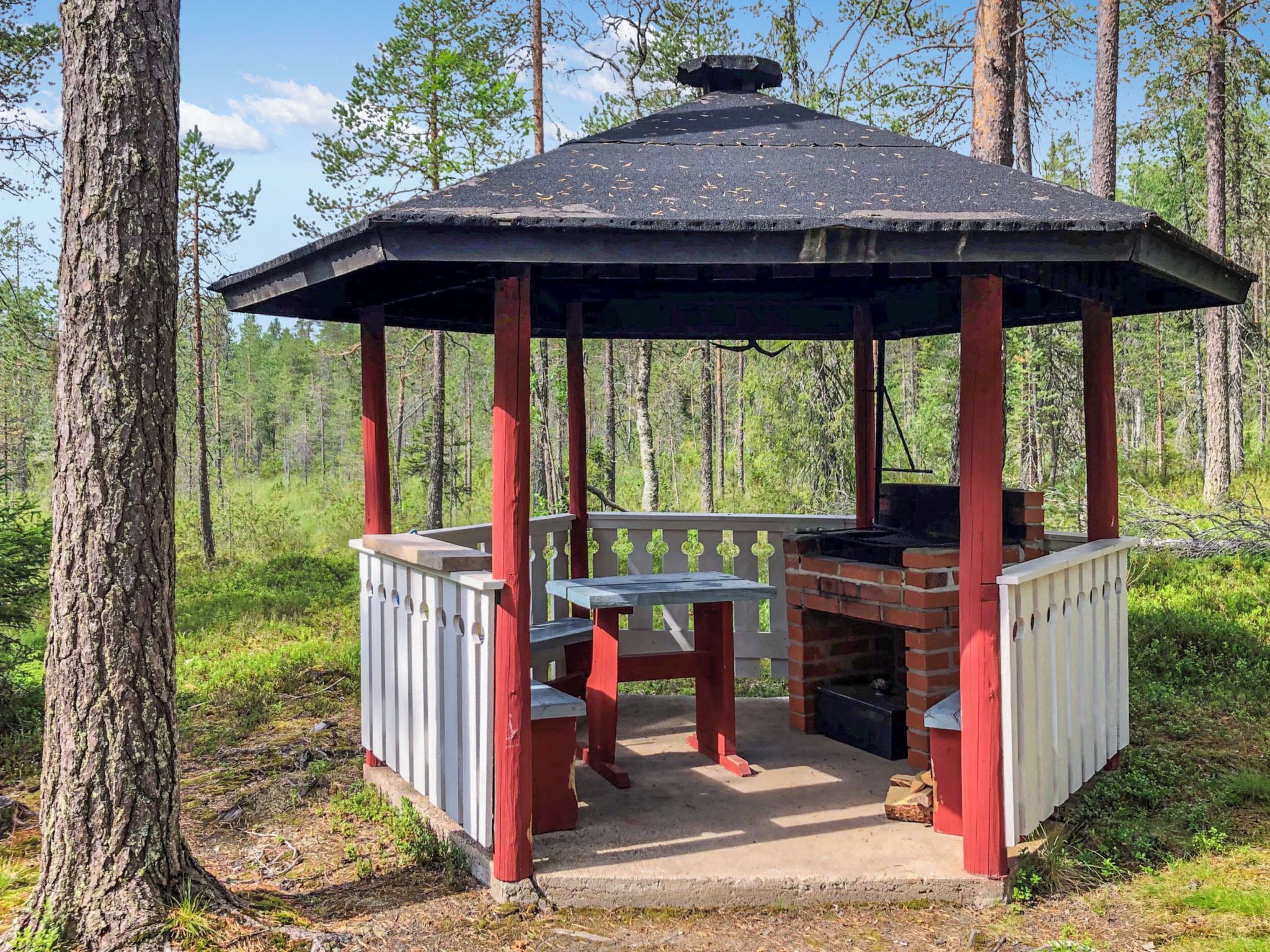A gazebo with a brick fireplace, wooden benches, and a metal roof in a forest setting.