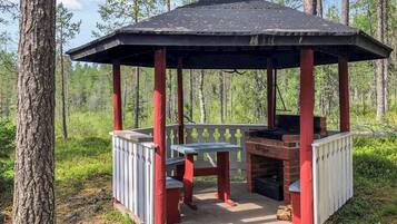 A gazebo with a brick fireplace, wooden benches, and a metal roof in a forest setting.