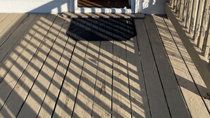 Eine Holzterrasse mit einer schwarzen Fußmatte, einer weißen Außenwand und einer Lampe.