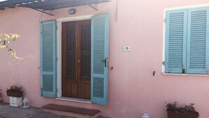 Un bâtiment rose avec une porte en bois et des volets bleus, une plaque d’immatriculation blanche et des plantes en pot.