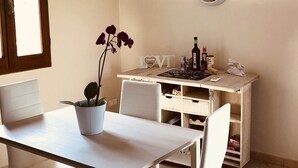 Un coin repas avec une table et des chaises, une plante en pot, une horloge murale et un petit espace cuisine comprenant un réfrigérateur et des bouteilles.