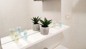 Ein Badezimmer mit weißen Fliesen, einer weißen Ablagefläche, zwei Aloe Vera Pflanzen und Toilettenartikeln, darunter Shampoo und Conditioner.