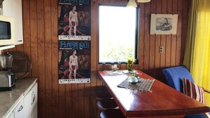A wooden-paneled room with a wooden dining table, chairs, a window with a view, and a poster on the wall. - Cabañas Mana Ora (Hanga Roa)