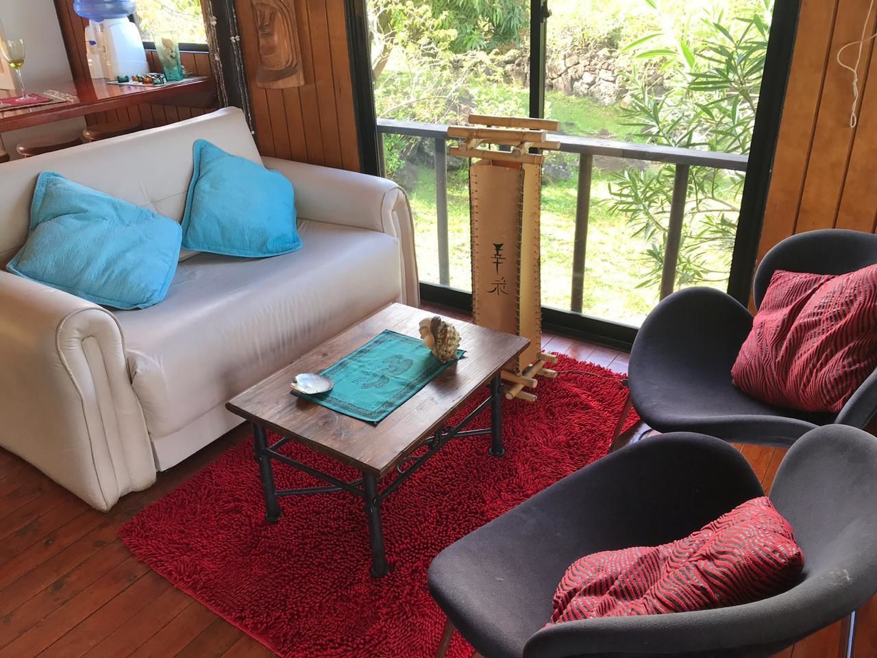 Ein gemütliches Wohnzimmer mit einem weißen Sofa, blauen Kissen, einem roten Teppich und Blick auf grüne Vegetation.