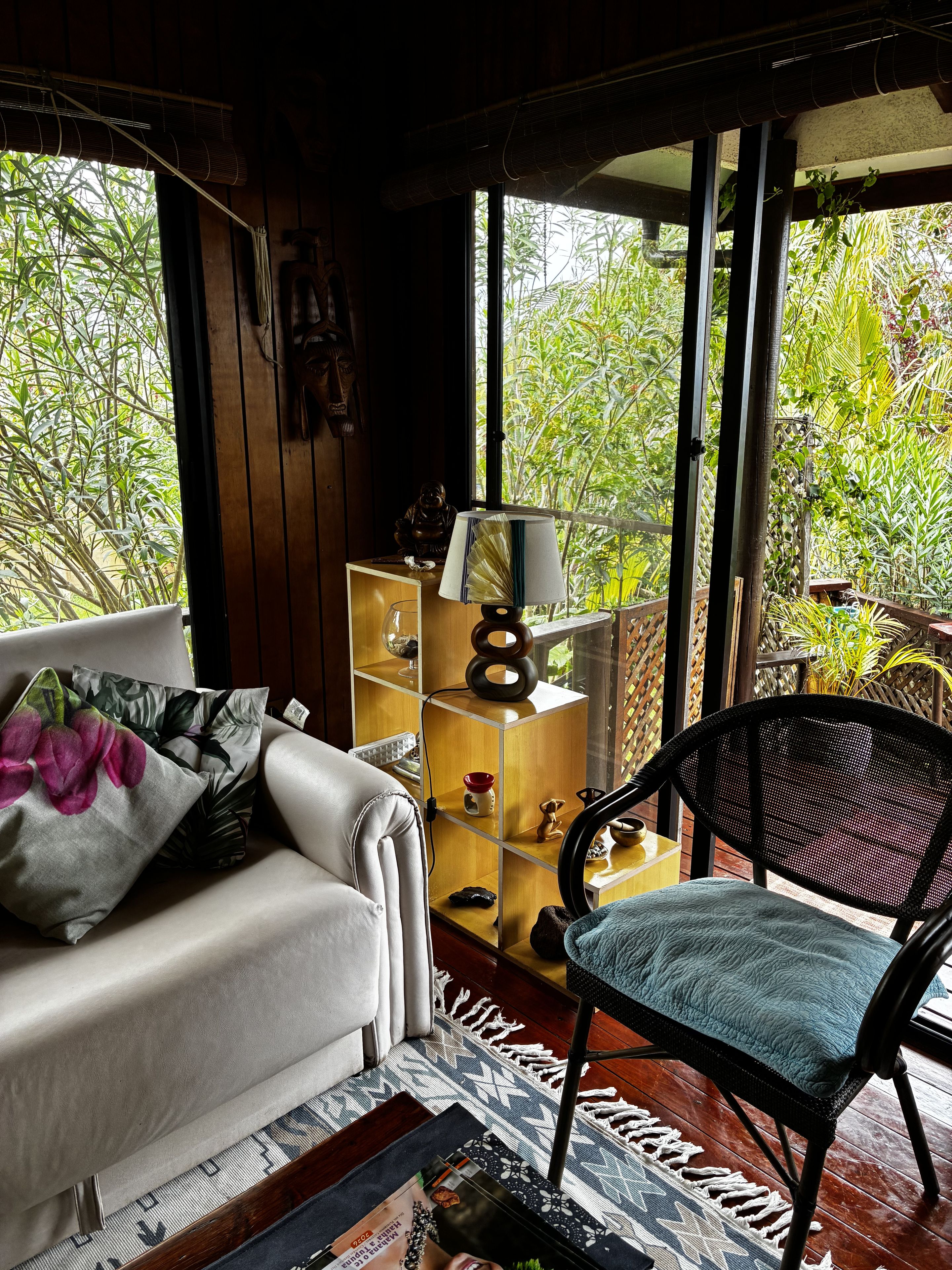 A cozy living room with a sofa, a chair, a wooden shelf with decorative items, and a view of lush greenery through large windows.
