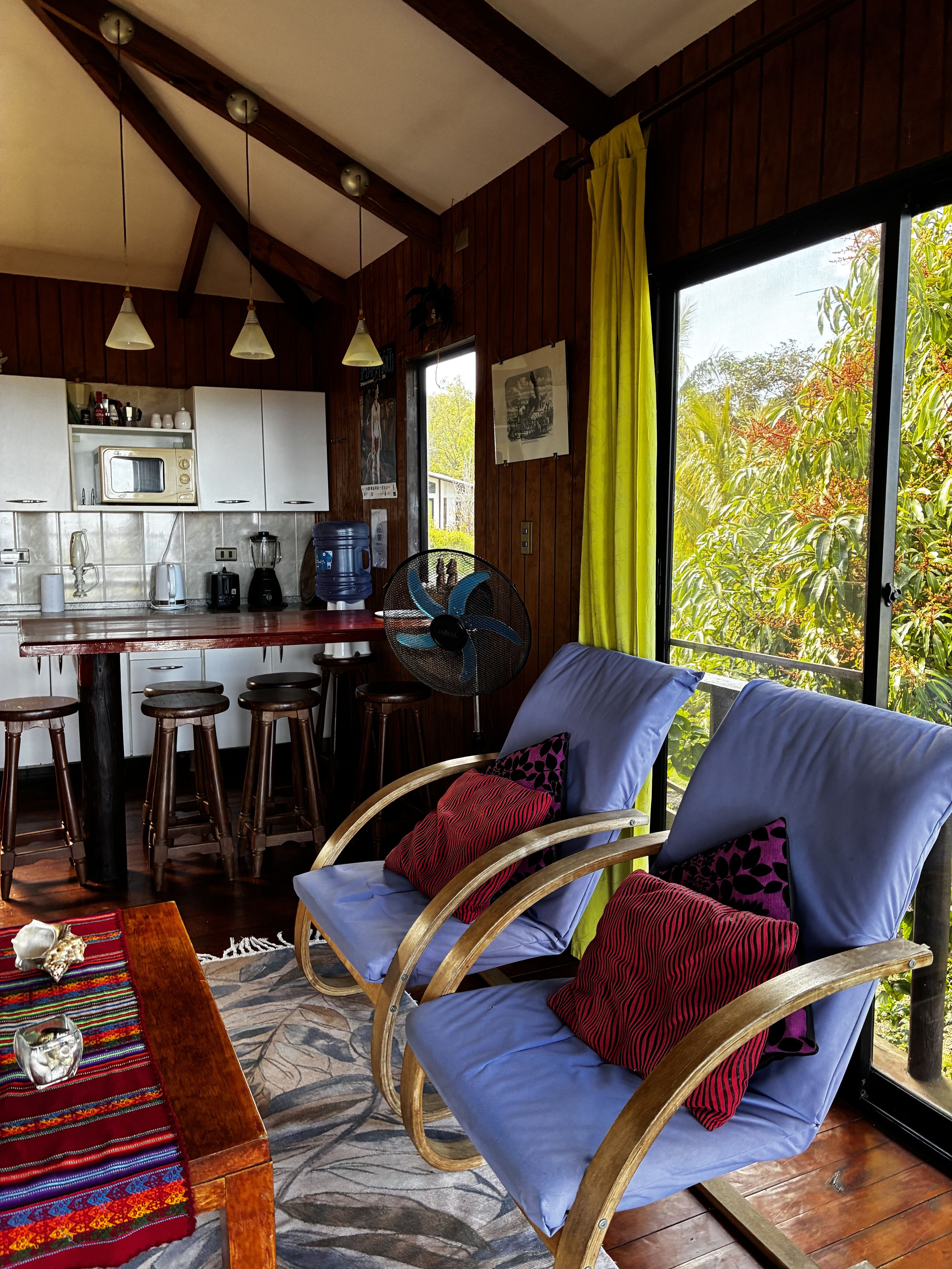 A room with a dining area, kitchen, and a large window with a view of greenery.