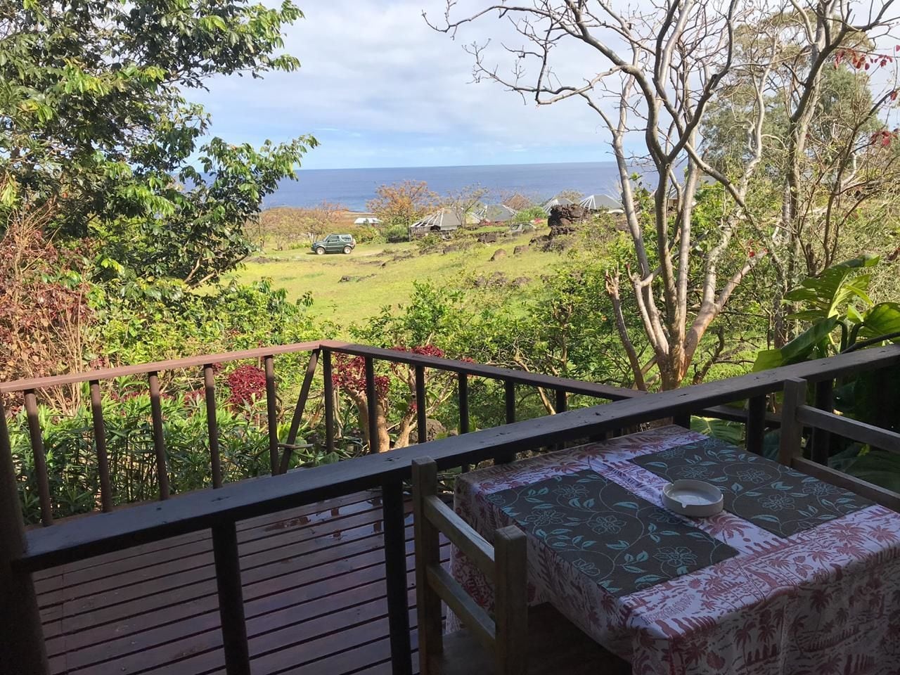 Un terrazzo in legno con tavolo e sedie, affacciato su un paesaggio rigoglioso e vista sull'oceano e case in lontananza.
