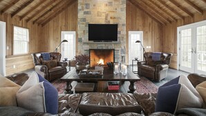 A cozy log cabin interior with a stone fireplace, leather furniture, and a large flat-screen TV mounted above the hearth.