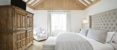 Un dormitorio con una cama grande, un armario de madera, un sillón blanco y una ventana con cortinas.